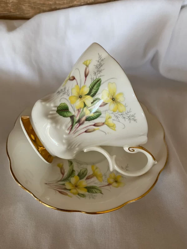 Royal Albert tea cups yellow flowers, green petals on white backdrop and gold trim cup atop saucer sideways